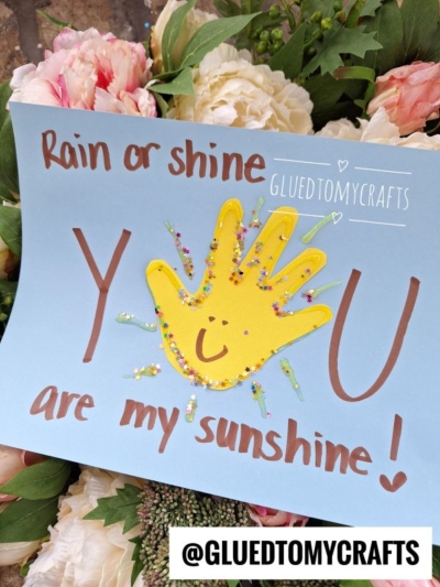 A handmade Easter Kid Craft card with "Rain or shine, you are my sunshine!" in red letters, a yellow handprint with a smiley face and glitter rays on blue paper, flowers in the background. @GLUEDTOMYCRAFTS is at the bottom.