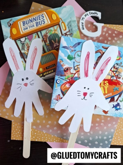 Two bunny-themed children's books rest on polka-dot paper. In front are Easter kid craft bunnies made from white paper handprints, with drawn faces and pink ears, attached to craft sticks. A @GLUEDTOMYCRAFTS label sits at the bottom.