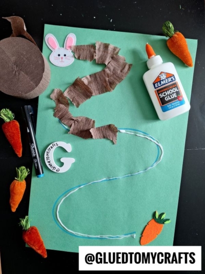 A green paper with a question mark outlined in yarn, partially covered in brown crepe paper. Surrounded by felt carrots, a bunny face, glue, scissors, and a pen—this Easter Kid Craft scene is finished with @GLUEDTOMYCRAFTS at the bottom.