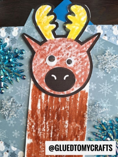 A craft reindeer made from paper and brown coloring, with googly eyes and yellow antlers, is displayed on a snowflake-patterned background—similar to a paper bag moose puppet. The bottom right corner has the text @GLUEDTOMYCRAFTS.