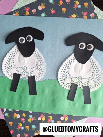 Two sheep made from paper doilies, black paper, and googly eyes are glued on blue and green paper to resemble a meadow. Surrounded by patterned craft papers, this charming Easter Kid Craft is signed @GLUEDTOMYCRAFTS at the bottom.