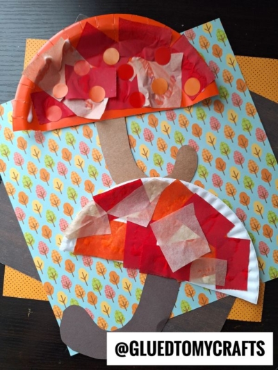 Two colorful paper mushrooms with tissue paper caps are glued onto autumn-themed leaf-patterned paper, resembling a whimsical forest of paper umbrella shapes. A watermark at the bottom reads @GLUETOMYCRAFTS.