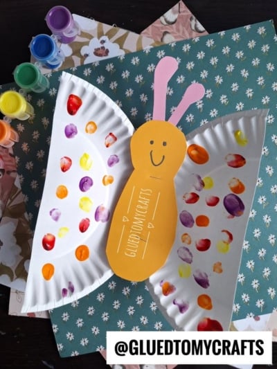 A colorful butterfly craft made from painted paper plates and orange paper, set on patterned paper—perfect as an Easter Kid Craft. Paint pots sit on the side. The handle @GLUEDTOMYCRAFTS is displayed at the bottom.