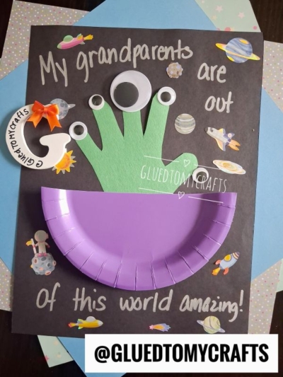 A kids craft card with “My grandparents are out of this world amazing!” in white writing, featuring a purple paper plate spaceship, a green handprint alien with googly eyes, and paper planets and stars on a black background—perfect for those looking for creative handprint gift ideas.