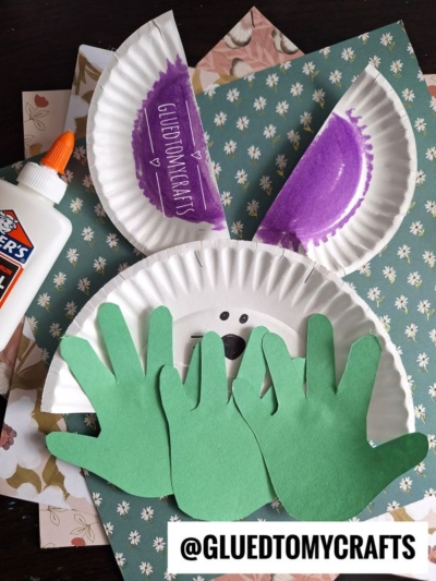 A fun Easter Kid Craft: a bunny made with paper plates, purple marker, green hand-shaped paper paws, black marker facial features, Elmer’s glue, and floral pattern paper. @GLUEDTOMYCRAFTS is written at the bottom.