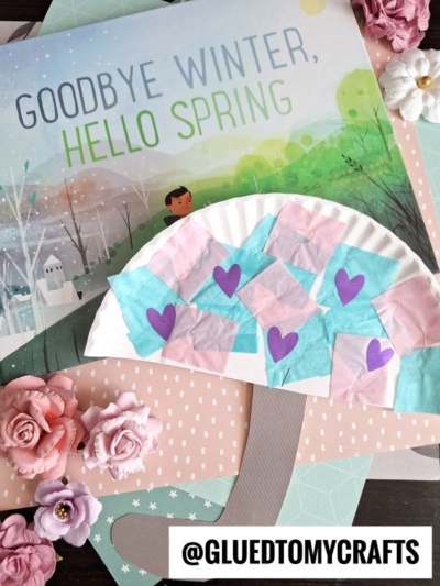A children’s book titled Goodbye Winter, Hello Spring is displayed with a spring-themed craft of a tree made from a paper plate, pastel tissue squares, and purple heart shapes. Pink artificial flowers and a paper plate umbrella decorate the scene.