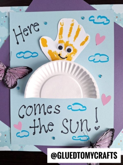A cheerful Easter Kid Craft featuring a semicircle paper plate, a yellow handprint sun with googly eyes, plus clouds, pink hearts, butterflies, and blue dots. The adorable craft displays the text: “Here comes the sun!”.