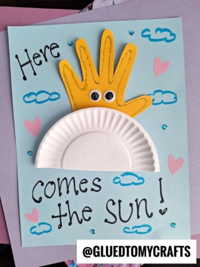 A handmade Easter Kid Craft card featuring a yellow handprint sun with googly eyes, blue clouds, pink hearts, and the words "Here comes the SUN!" Paper plate clouds complete this sweet creation. Labeled @GLUEDTOMYCRAFTS.