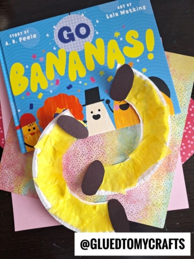 A children's book titled Go Bananas! sits on colorful paper, beside two banana crafts made from painted paper plate bananas with brown tips. The handle @GLUEDTOMYCRAFTS is printed at the bottom.
