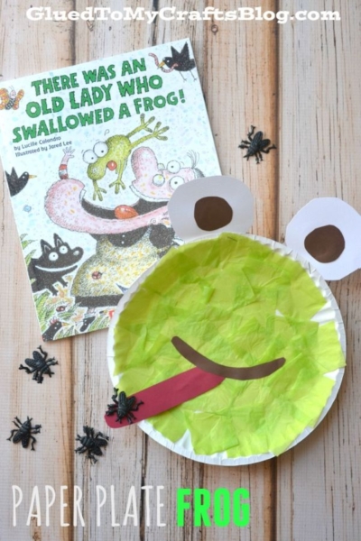 A children's book titled There Was an Old Lady Who Swallowed a Frog! is next to a colorful paper plate frog craft, surrounded by small plastic black spiders on a wooden surface.