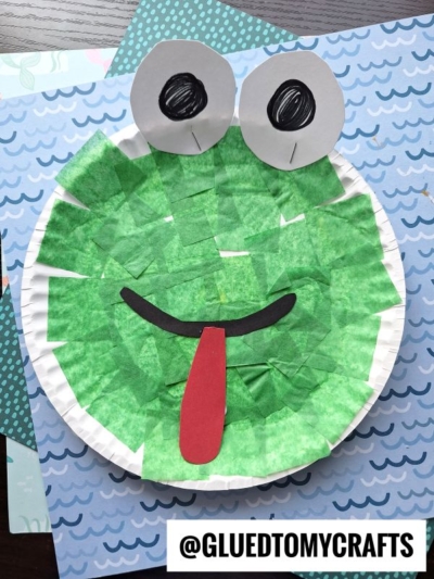 A cute paper plate frog craft made with green tissue paper squares, big white eyes, a black smiling mouth, and a red tongue. Set against blue patterned paper. Text: @GLUETOMYCRAFTS.