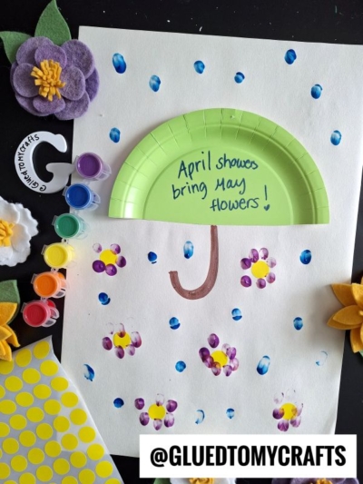 A spring-themed Easter kid craft featuring a green paper plate cut into an umbrella shape, painted raindrops, and finger-painted flowers, surrounded by felt flowers, paint pots, yellow stickers, and a @GLUEDTOMYCRAFTS label.