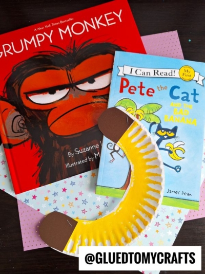 Two children’s books, “Grumpy Monkey” and “Pete the Cat and the Bad Banana,” are on a table with a yellow paper plate banana craft. The handle @gluedtomycrafts appears at the bottom.