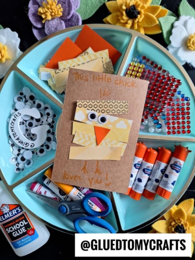 A craft tray with supplies like glue, scissors, googly eyes, markers, felt flowers, and colored paper strips for a fun Easter Kid Craft. Includes a card featuring a chick made from yellow paper strips, googly eyes, and an orange beak. Text: “@GLUEDTOMYCRAFTS”.