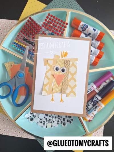 A handmade Easter Kid Craft card featuring a paper chick made of patterned strips, googly eyes, and an orange beak, displayed on a tray with craft supplies like glue, scissors, markers, and gem stickers.