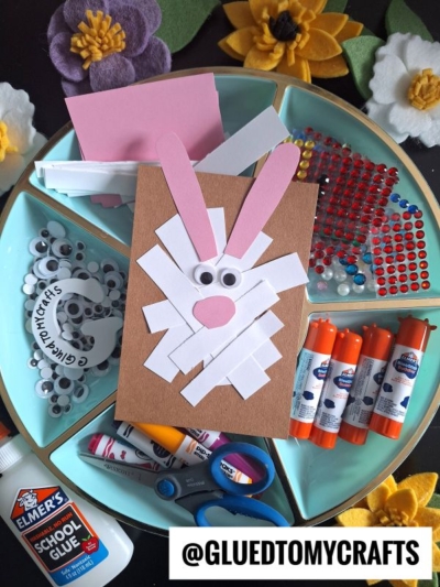 A round tray holds craft supplies, including glue sticks, scissors, googly eyes, colored paper, rhinestones, and felt flowers. At the center is a handmade bunny card—an adorable Easter kid craft made from brown paper and white strips.