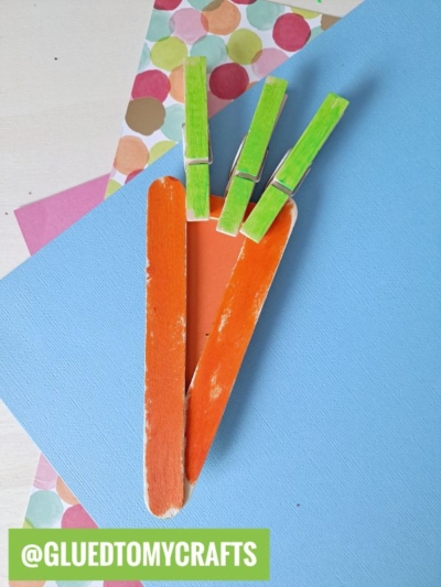 Three orange popsicle sticks arranged in a carrot shape with three green clothespins as leaves create a fun Easter Kid Craft, placed on blue and colorful polka dot paper. @GLUEDTOMYCRAFTS is written at the bottom.