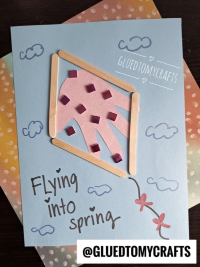 A handmade Easter kid craft featuring a pink handprint kite with popsicle sticks, purple squares, and cloud doodles on blue paper. The text reads “Flying into spring” and “@GLUEDTOMYCRAFTS.”.