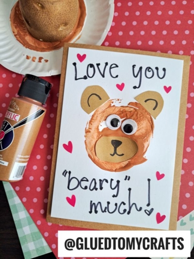 A handmade Potato Stamped Bear Card features a bear face made from brown paint and googly eyes, surrounded by pink hearts with the message "Love you beary much!" Art supplies and a painted potato sit nearby. Handle: @GLUEDTOMYCRAFTS.