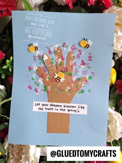 A blue paper background displays a brown paper handprint tree with pink, green, and white sequins as blossoms—an adorable Easter Kid Craft. Smiling bee stickers buzz around. A note reads, Let your dreams blossom like the trees in the spring!.