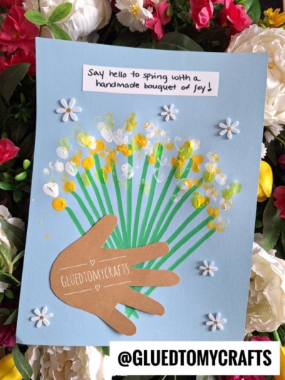 A charming Easter Kid Craft project features a brown paper handprint as flower stems, with yellow and white painted dots as flowers on blue paper. White paper flowers and the text Say hello to spring with a handmade bouquet of joy! are included.