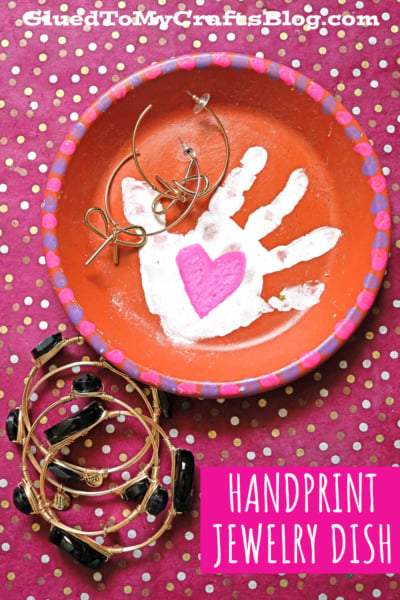 A homemade terra cotta jewelry plate features a painted white handprint and a pink heart in the center, holding earrings. The orange dish with a pink edge rests on a pink polka-dotted background with bracelets beside it.