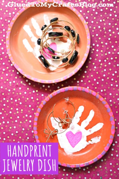 Two handmade terra cotta jewelry plates painted orange, each featuring a white handprint with a pink heart in the center. The dishes hold jewelry and are placed on a pink polka dot background. Text reads HANDPRINT JEWELRY DISH.