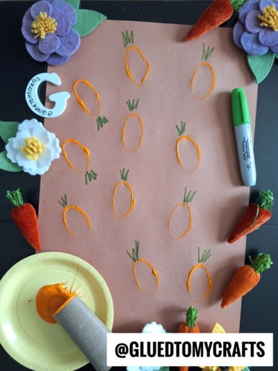 A charming Easter Kid Craft featuring a brown paper with orange and green stamped carrot shapes, surrounded by felt flowers and carrots. A green marker, orange paint, and stamp sit nearby. The handle @GLUEDTOMYCRAFTS is at the bottom.