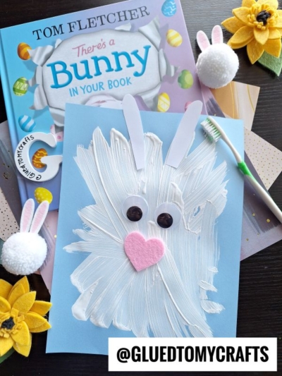 A children’s book titled “There’s a Bunny in Your Book” sits next to an adorable Easter kid craft: a bunny made from white paint, paper ears, button eyes, and a pink felt nose. Pom-pom bunnies, yellow fabric sunflowers, and a toothbrush are nearby.