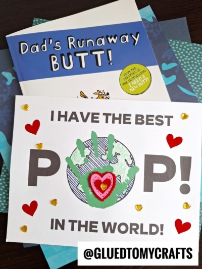 A handmade Fathers Day card with “Handprint Best Pop in the World!” and a green handprint forming the “O,” decorated with hearts, sits atop the book “Dad’s Runaway Butt.”.