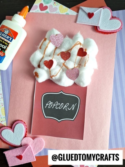 A handmade Valentine’s card with cotton ball popcorn, heart-shaped embellishments, and a “popcorn” label. Felt hearts, envelopes, and a bottle of glue are nearby. Perfect for a fun popcorn kid craft! Image tagged @GLUEDTOMYCRAFTS.