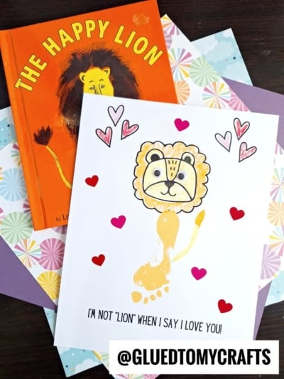 A children’s book titled The Happy Lion and a handmade lion card with hearts are arranged on colorful paper. The card features a charming lion footprint keepsake, with the lion drawn around a footprint and the text, I’M NOT LION WHEN I SAY I LOVE YOU!.