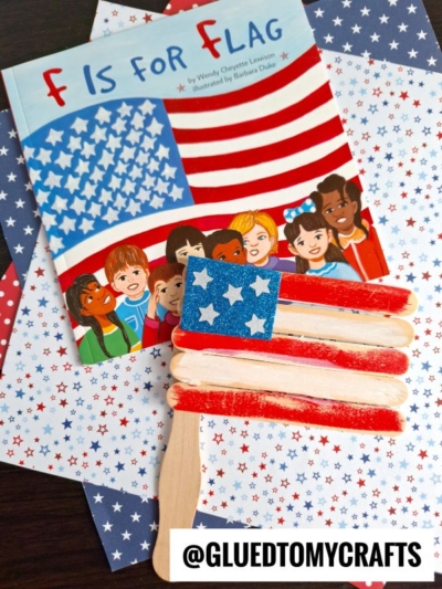 A children’s book titled F Is for Flag sits on star-patterned paper, next to a flag kid craft—popsicle stick flags decorated in red, white, and blue with white stars. @GLUEDTOMYCRAFTS is written at the bottom.