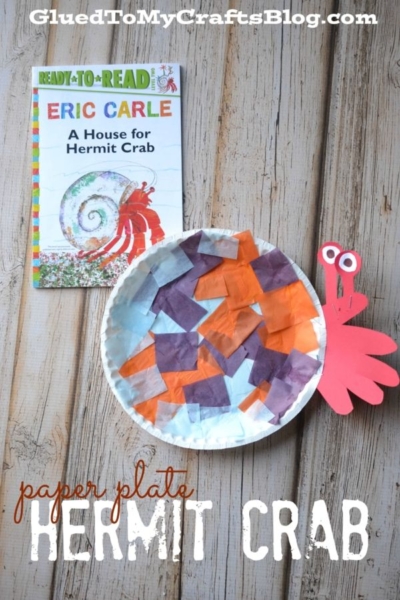 A children’s book titled A House for Hermit Crab and a colorful paper plate hermit crab craft—made with tissue paper squares and bright red paper claws—are displayed on a wooden surface.