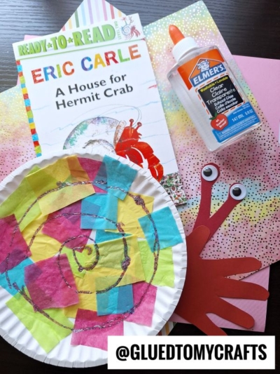 Children’s craft supplies with “A House for Hermit Crab” book, a paper plate hermit crab decorated with colorful tissue pieces, glue bottle, construction paper crab, and patterned paper sheets. @GLUEDTOMYCRAFTS at the bottom.