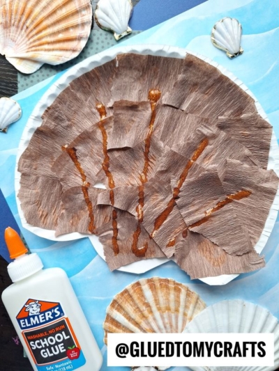 A paper plate seashell kid craft decorated with brown crepe paper and orange glue lines, surrounded by sea shells and a bottle of Elmer’s glue on a blue surface. @GLUEDTOMYCRAFTS is written at the bottom.