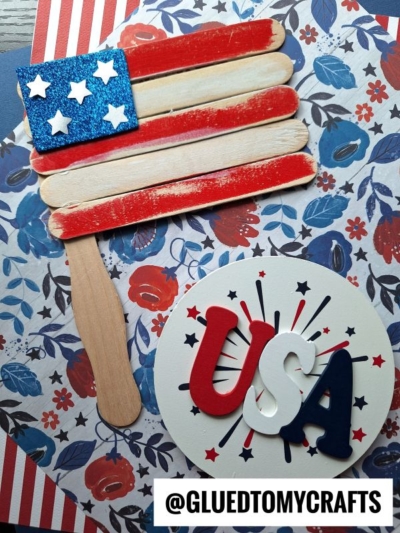 A patriotic flag kid craft made from popsicle stick flags painted as an American flag, with a blue glitter corner and white stars, displayed on floral paper and topped with a round card reading USA in red, white, and blue. @GLUEDTOMYCRAFTS.