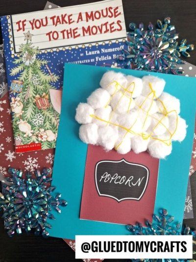 A children's book, If You Take a Mouse to the Movies, sits next to a popcorn kid craft made with cotton ball popcorn and yellow string, labeled Popcorn. Snowflake decorations and a @GLUEDTOMYCRAFTS tag are visible nearby.