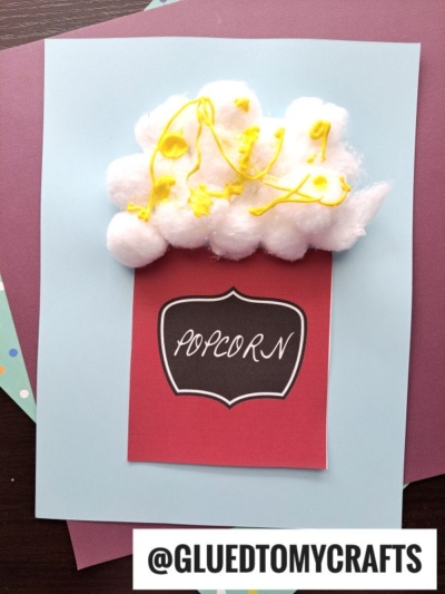A fun popcorn kid craft features cotton ball popcorn with yellow paint for butter, glued above a red paper container labeled POPCORN on a blue background. @GLUEDTOMYCRAFTS is written at the bottom.