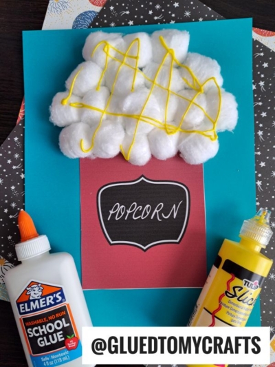 A blue paper displays a popcorn kid craft made with cotton ball popcorn and yellow puffy paint for kernels, labeled “POPCORN.” School glue and yellow paint bottles sit nearby. The handle @GLUEDTOMYCRAFTS is shown at the bottom.