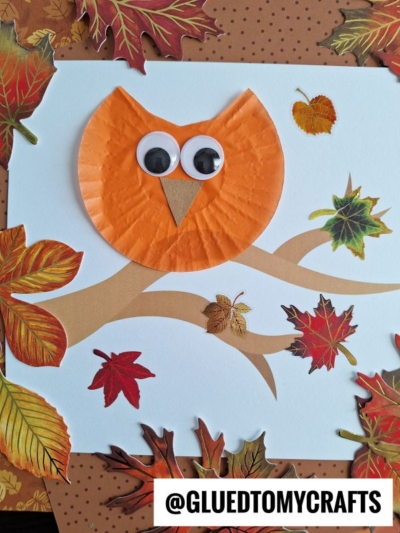 A fall-themed craft featuring a cupcake liner owl with large googly eyes and a beak, perched on a paper tree branch and surrounded by colorful autumn leaves. @gluedtomycrafts is written at the bottom.