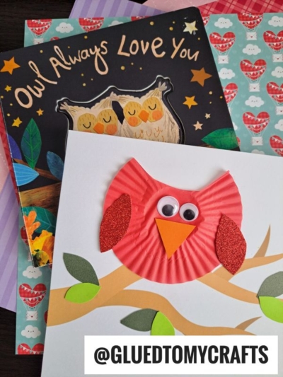 A handmade cupcake liner owl card with googly eyes and red glitter wings sits on top of an “Owl Always Love You” book and colorful, patterned paper. The username @GLUEDTOMYCRAFTS is visible at the bottom.