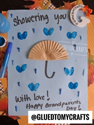 A blue card features a charming cupcake liner umbrella craft, with a brown cupcake liner as the umbrella, blue painted raindrops and hearts, and the words “Showering you with love! Happy Grandparents Day!” Art supplies and leaves are nearby.