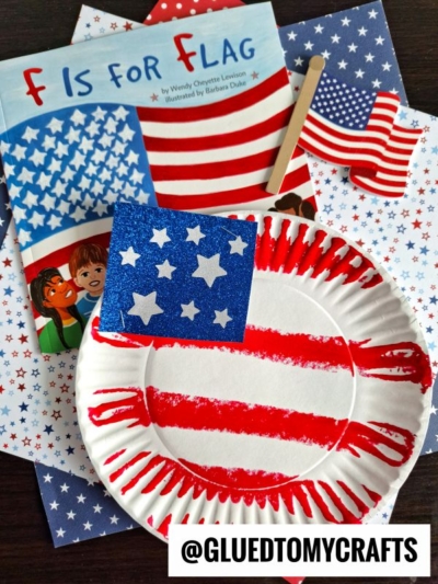 A paper plate flag decorated with red paint and a blue square with white stars sits next to the book F Is for Flag, star-patterned paper, and another fun flag craft for kids. @GLUEDTOMYCRAFTS text appears at the bottom.