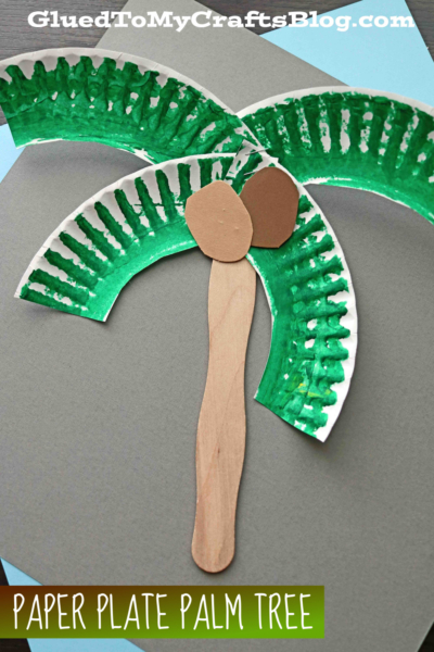 A palm tree paper plate craft made from painted paper plate pieces for leaves and wooden craft sticks for the trunk, with two brown paper coconuts, displayed on gray paper.