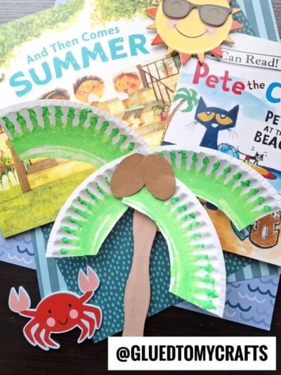 A summer-themed flat lay featuring children’s books, a palm tree paper plate craft with green painted leaves and brown coconuts, a smiling sun, and a red paper crab on a striped background.