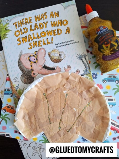 A children’s book titled There Was an Old Lady Who Swallowed a Shell! is next to a seashell kid craft with glue, glitter, a bottle of gold glitter glue, and craft supplies on a decorated surface. Text: @GLUEDTOMYCRAFTS.
