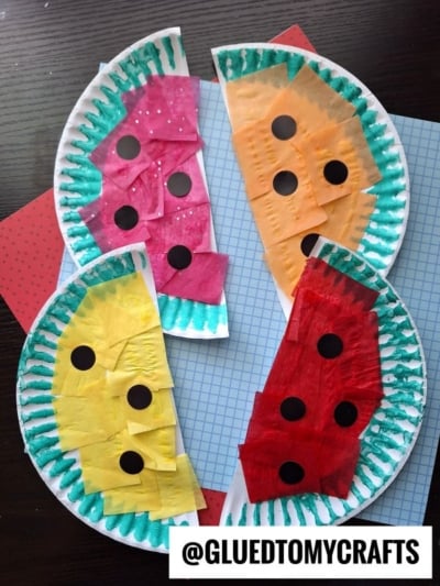 Three paper plate watermelon crafts decorated with red, yellow, and orange tissue paper, green marker for the rind, and black dots for seeds are arranged on a patterned background. @GLUEDTOMYCRAFTS is at the bottom.