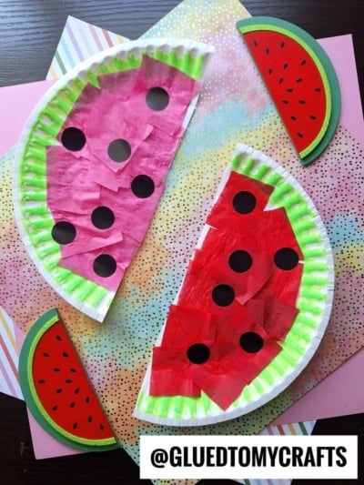 Two paper plate watermelon slices are crafted with green and pink tissue paper and black paper seeds, displayed on vibrant patterned paper with festive watermelon-themed decorations. @GLUEDTOMYCRAFTS text at the bottom.