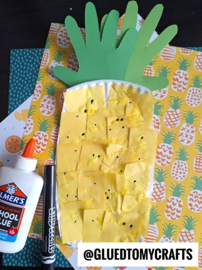 A handmade paper pineapple craft made from yellow paper squares, black marker dots, and green paper leaves sits beside a glue bottle and marker. Colorful pineapple-patterned paper is in the background.